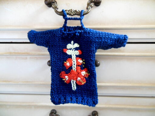 Hand knit minature jumper ornament in navy blue with an embroidered gladioli flower design shown attached to a handle on a chest of draws.